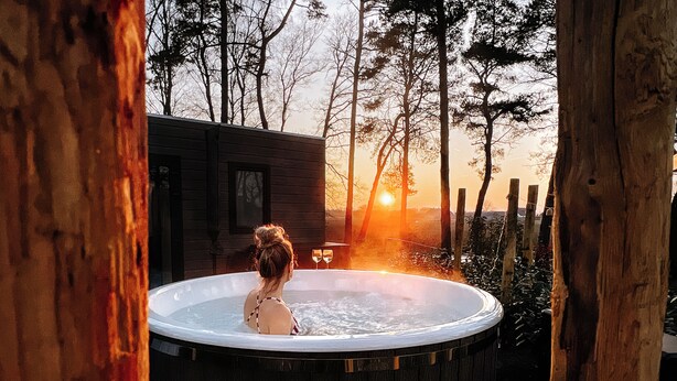 Une personne se détend dans un bain à remous, avec une vue sur le coucher de soleil, entourée d’arbres et d’un chalet en bois.