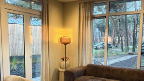 Un salon moderne avec un canapé, un fauteuil et une table basse, de grandes fenêtres avec des rideaux et une lampe.