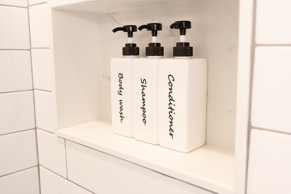 Une étagère de salle de bain avec trois flacons blancs étiquetés « Gel douche », « Shampoing » et « Après-shampoing ».