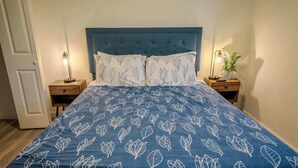 A bedroom with a blue bedspread featuring a leaf pattern, a tufted headboard, two bedside tables with lamps, and a potted plant on one table. - Stay Roanoke | Charming 1BR Near Hospital & City (Roanoke)