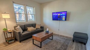 A living room with a grey sofa, a wooden coffee table, a grey ottoman, a TV mounted on the wall, and a window with a view of buildings. - Stay Roanoke | Charming 1BR Near Hospital & City (Roanoke)
