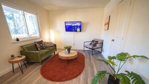 Ein Wohnzimmer mit einem grünen Sofa, einem roten Teppich, einem runden Couchtisch, einem an der Wand montierten Fernseher und einer Topfpflanze.