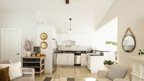 A spacious living room with a sofa, a coffee table, and a fireplace. There is a kitchen in the background with white cabinets and a dining area.