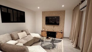 A modern living room with a sectional sofa, a glass coffee table, a flat-screen TV mounted on a textured wall, and a wooden cabinet below. - Spacious house near the lake - 6 people (Saint-Épain)