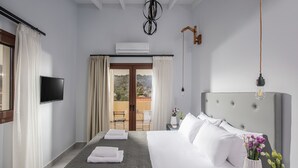 A modern hotel room with a large bed, a flat-screen TV, a balcony with curtains, and a view of the hills. - Iris Villa 3 (Archanes-Asterousia)