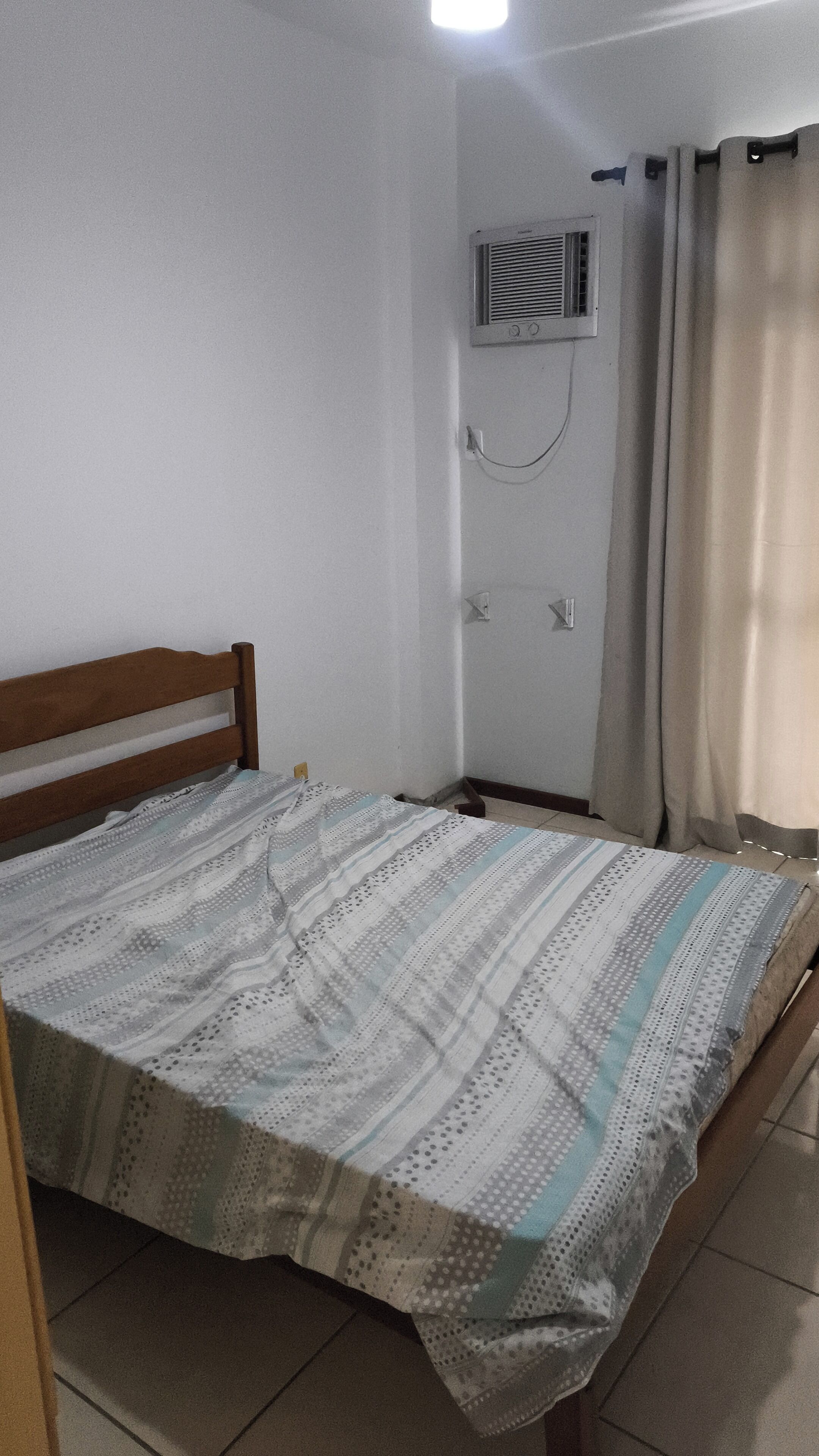 Ein kleines Zimmer mit einem Einzelbett, einem Holzkopfende, einer gemusterten Tagesdecke, einer Wandklimaanlage und einem Vorhang.