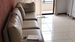 Ein beiges Sofa mit Kissen und einem Notizbuch, ein gefliester Boden, ein Fenster mit Jalousien und eine Tür, die nach draußen führt.