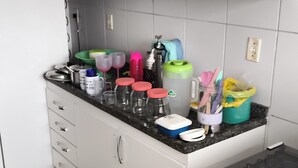 Eine Küche mit weißen Schränken, einer schwarzen Arbeitsplatte und einem Herd. Auf der Arbeitsplatte liegen verschiedene Küchenutensilien, darunter eine grüne Thermoskanne, eine pinkfarbene Flasche und ein blauer Behälter.