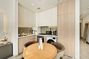 A modern kitchen with white cabinets, a round wooden table, and a washing machine. - Curated High-Floor 1BR / Water View / Peninsula 1 (Dubai)