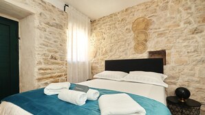 A room with a stone wall, a bed with a blue bedspread, white pillows, and a black headboard. There are towels on the bed and a small round table with a lamp. - Holiday Home Tranquilla with heated pool (Blato)