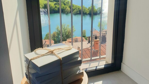 A balcony with a view of a lake and mountains, a stack of books on a small table, and a window with a metal railing. - The little door. Your lake view home. (Barrea)