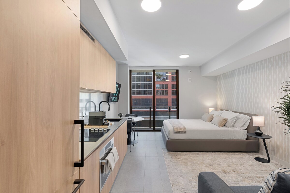 A modern, well-lit bedroom with a large bed, a bedside table with a lamp, and a view of a building through a sliding door.