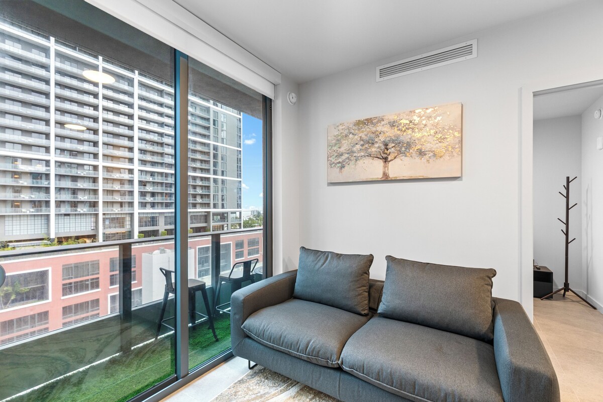 A modern living room with a large window, a grey sofa, and a painting of a tree.