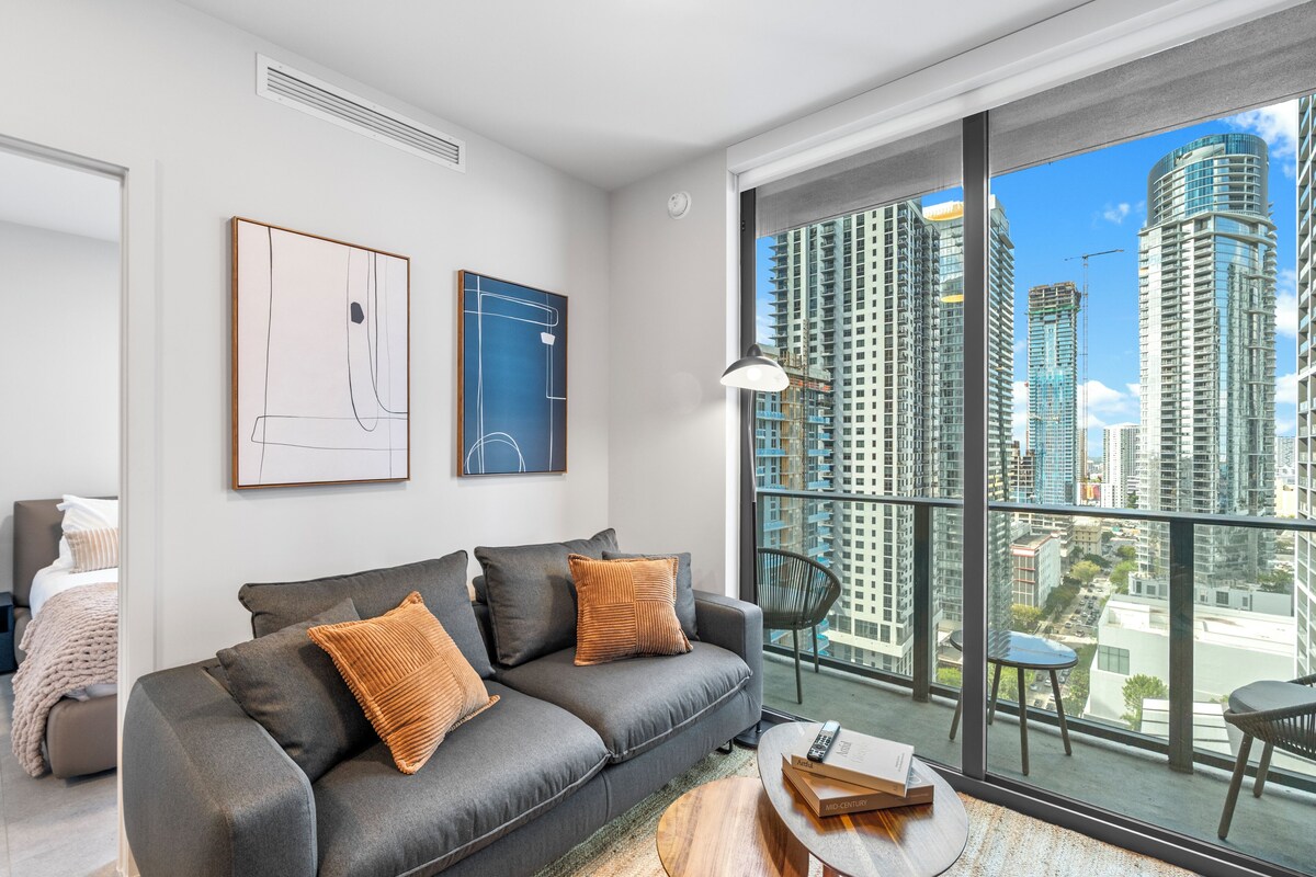 A modern living room with a grey sofa, two cushions, a coffee table, and a large window with a city view.