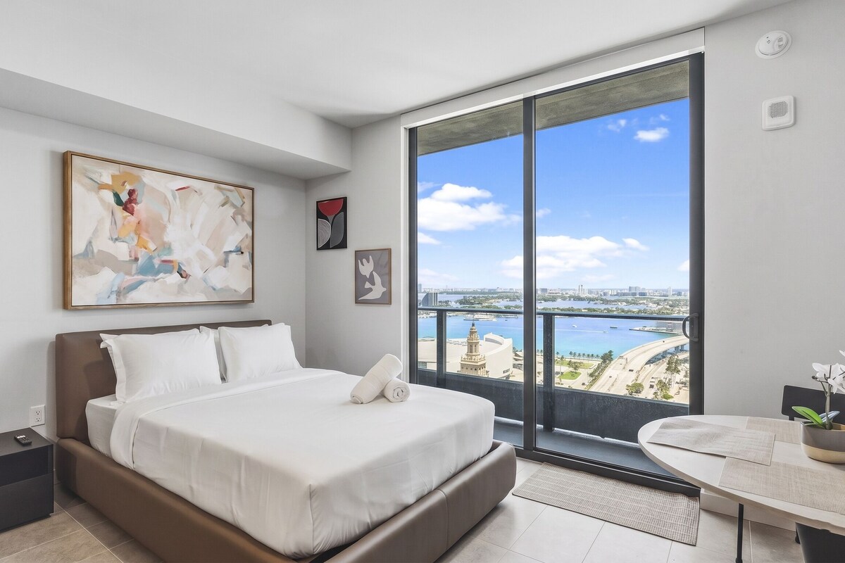 A modern bedroom with a large bed, a view of the city, and artwork on the wall.