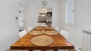 Un coin repas avec une table en bois, des chaises blanches, des sets de table en fibres tressées, une horloge et, au fond, une cuisine.