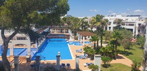 Ein großer Pool mit einer Brücke, umgeben von Palmen und Liegestühlen, in einem Resort mit modernen Gebäuden.