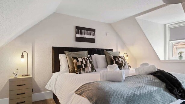A bedroom with a bed, pillows, a bedside table, a lamp, and a window. - Leo Apartment - 4 Bedroom Flat in Nairn (Nairn)