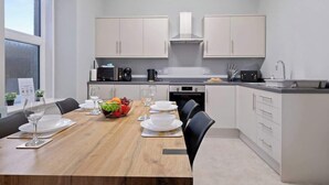 A modern kitchen with white cabinets, a wooden dining table set with plates and glasses, and a fruit bowl. - Leo Apartment - 4 Bedroom Flat in Nairn (Nairn)