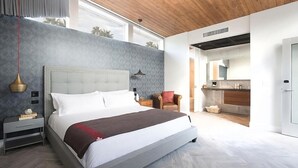 A modern bedroom with a large bed, a nightstand, a chair, and a view of the kitchen.
