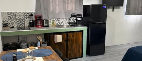 A compact kitchen with a refrigerator, coffee maker, and microwave. There is a dining table with placemats and a chair.
