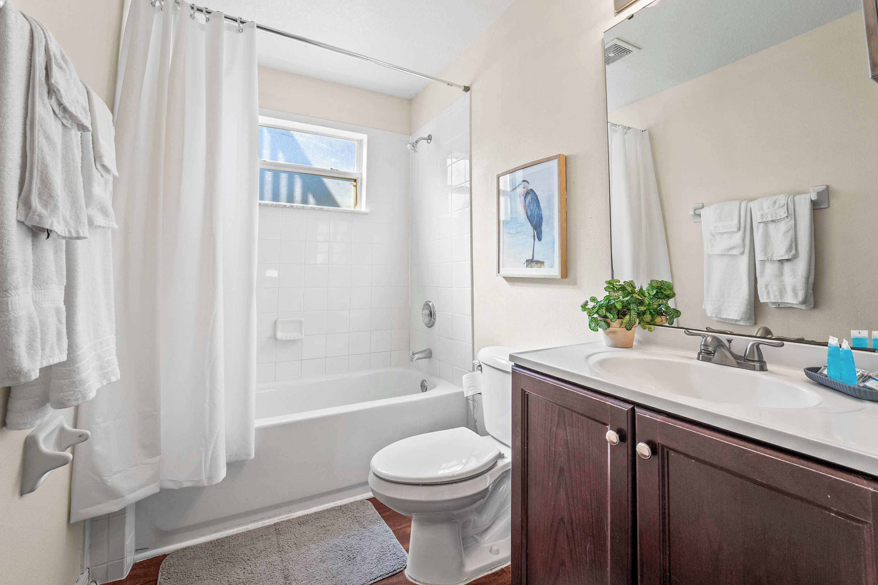 Un bagno con vasca, servizi igienici e lavandino. Si vede una finestra, un vaso con una pianta e un quadro appeso al muro.