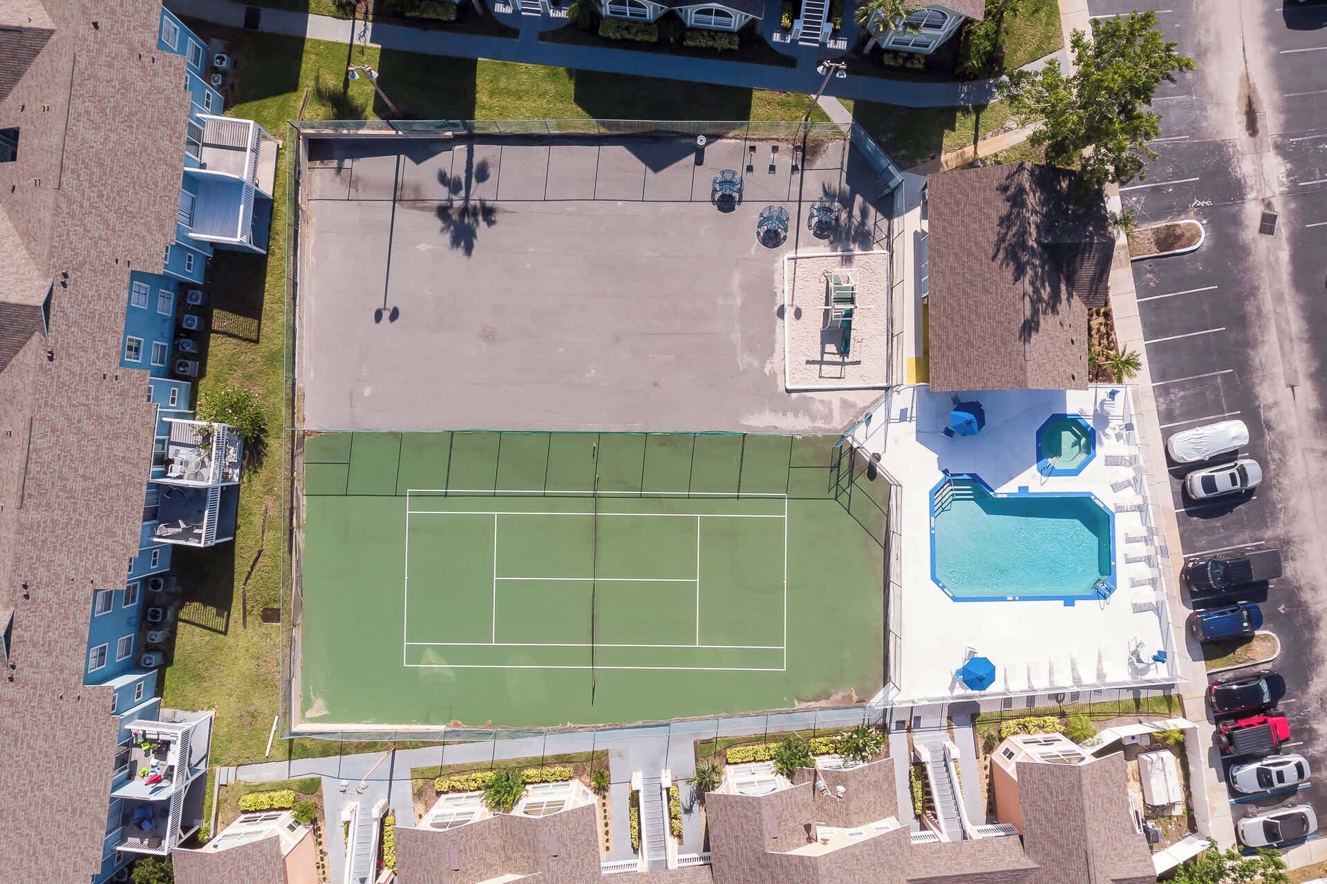 Vue aérienne d’un quartier résidentiel avec un court de tennis, une piscine et plusieurs voitures garées.