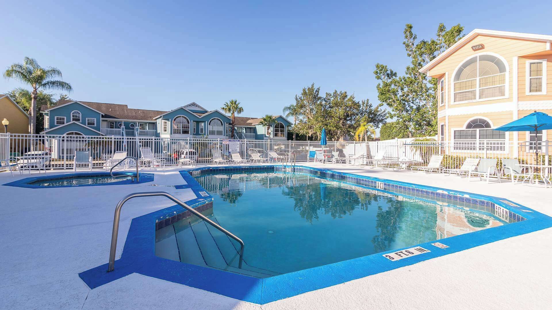 Une piscine dont le bord est bleu, entourée de chaises longues blanches et de parasols, avec des bâtiments en arrière-plan.