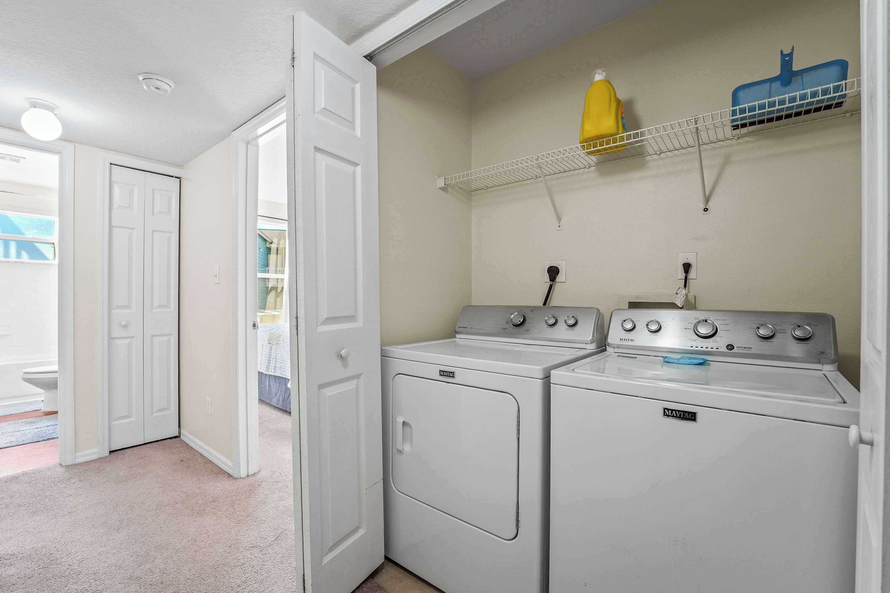 A laundry room with two white front-loading washers and dryers, a white door, and a shelf with a yellow bottle and a blue container.