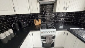 Une cuisine avec des meubles blancs, un dosseret de carreaux noirs, une cuisinière à gaz et une machine à café.