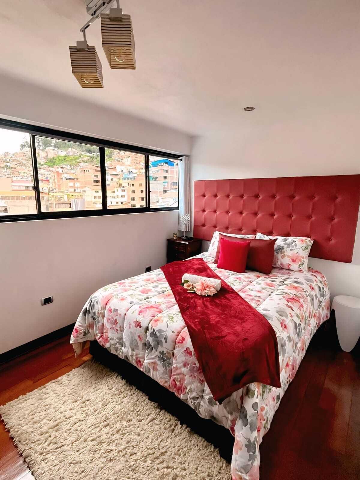 A bedroom with a large window, a red headboard, and a floral bedspread.