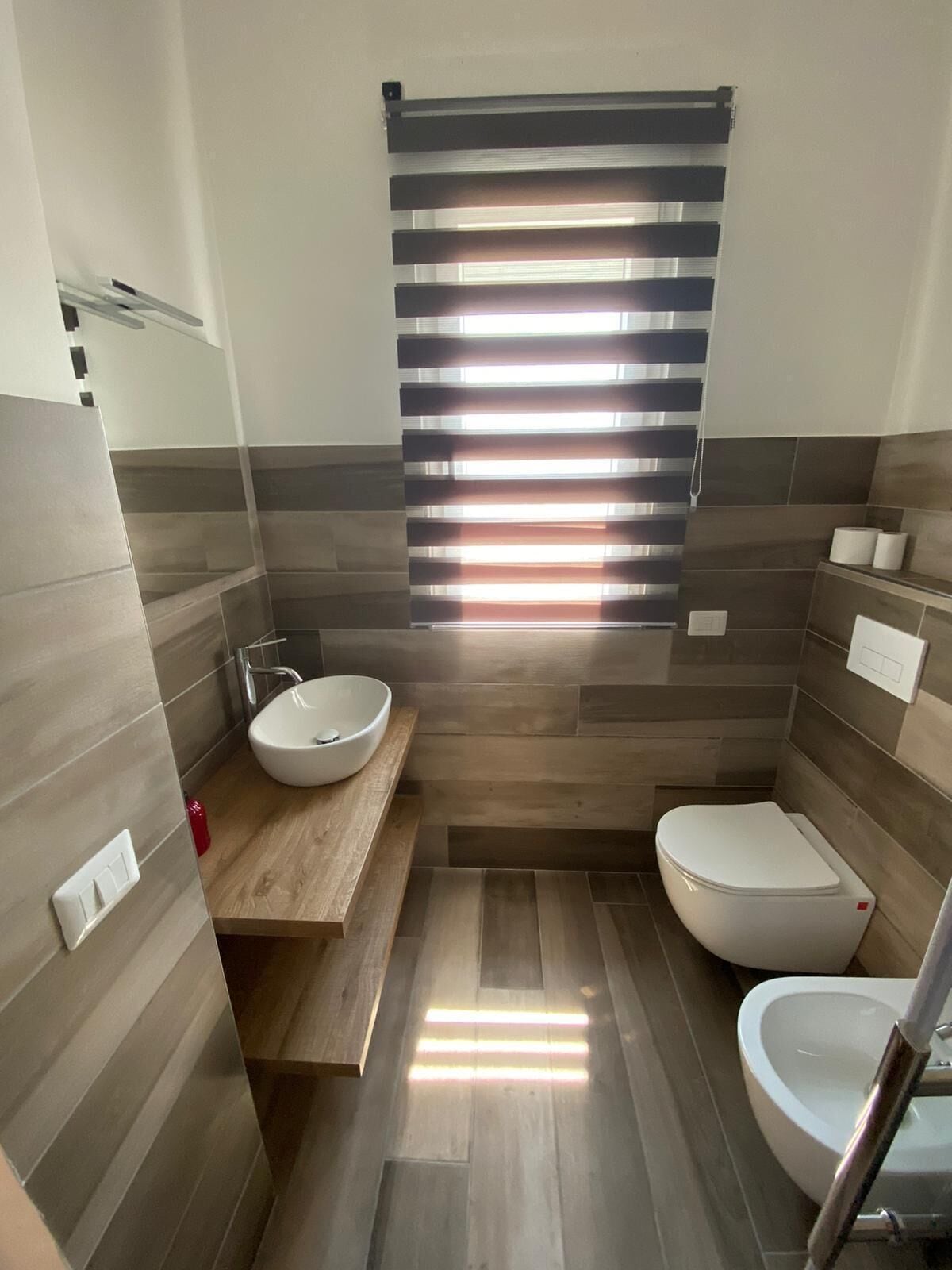 Un baño moderno con tocador de madera, un lavabo blanco, un inodoro y una ventana con persianas horizontales.