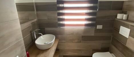 Un baño moderno con tocador de madera, un lavabo blanco, un inodoro y una ventana con persianas horizontales.