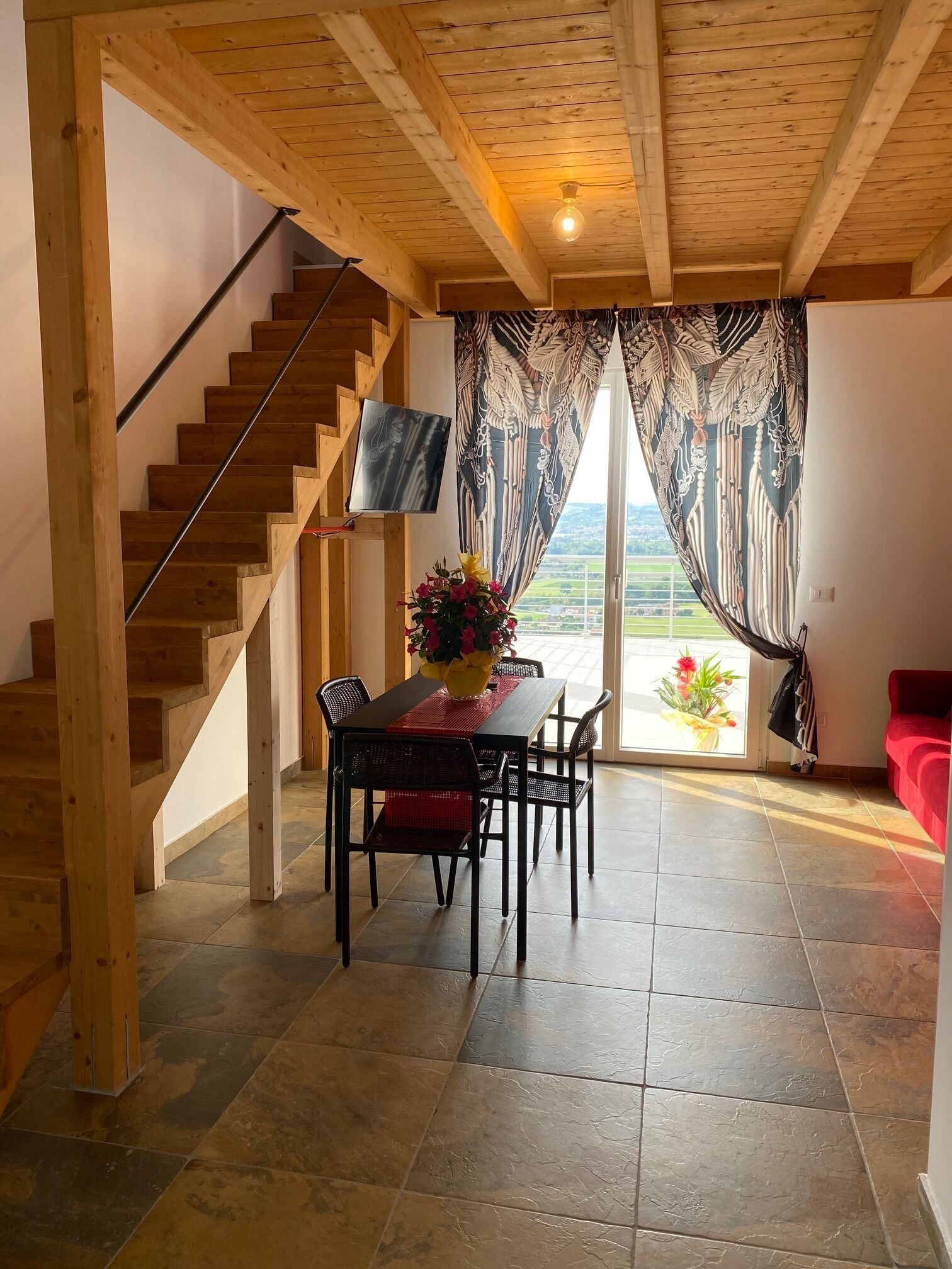 Un escalier en bois menant à un balcon avec vue, un coin repas avec une table et des chaises, et un canapé rouge.