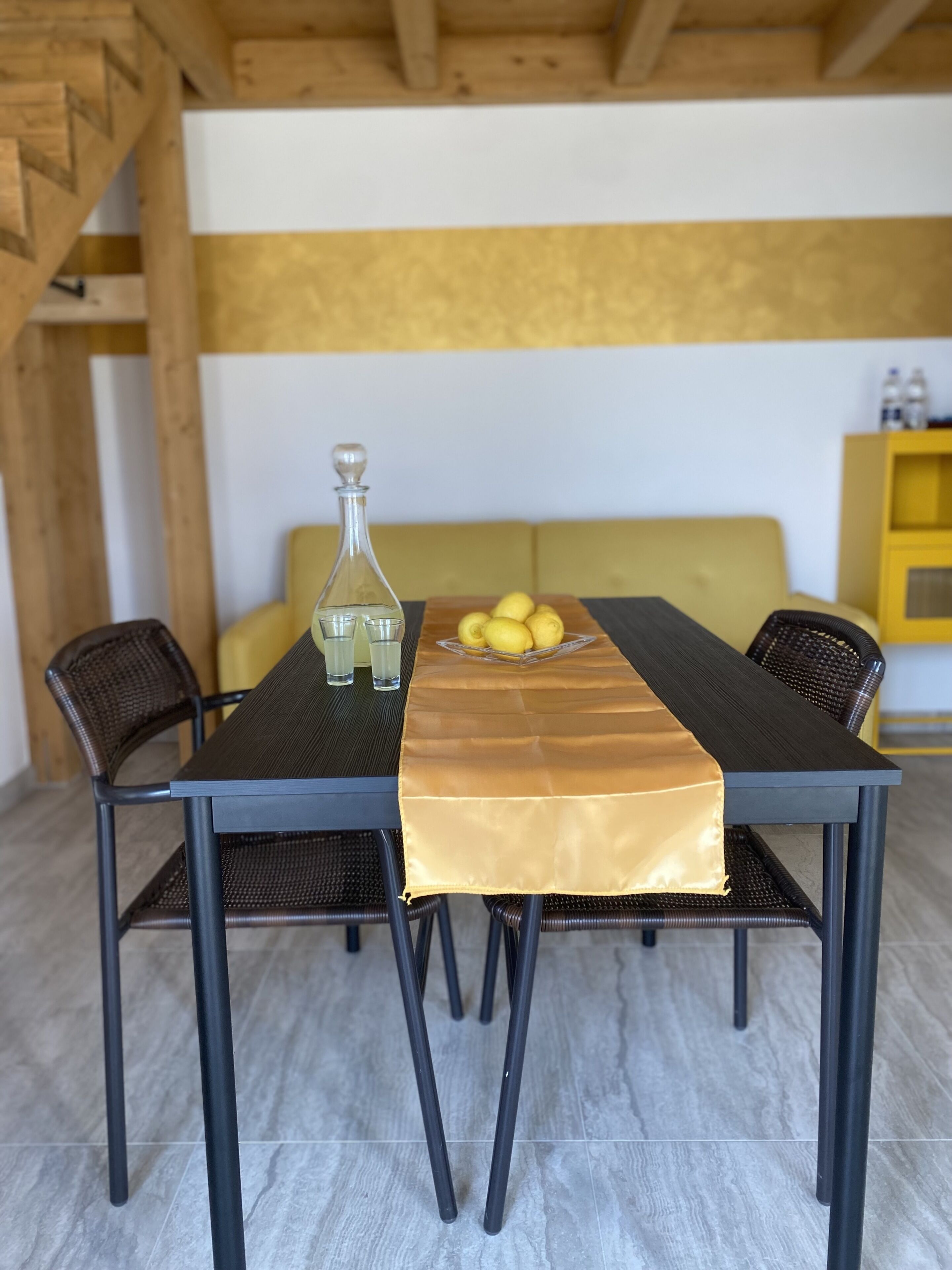 A dining table with a bottle and lemons, surrounded by wicker chairs, in a room with a wooden ceiling and a yellow wall.