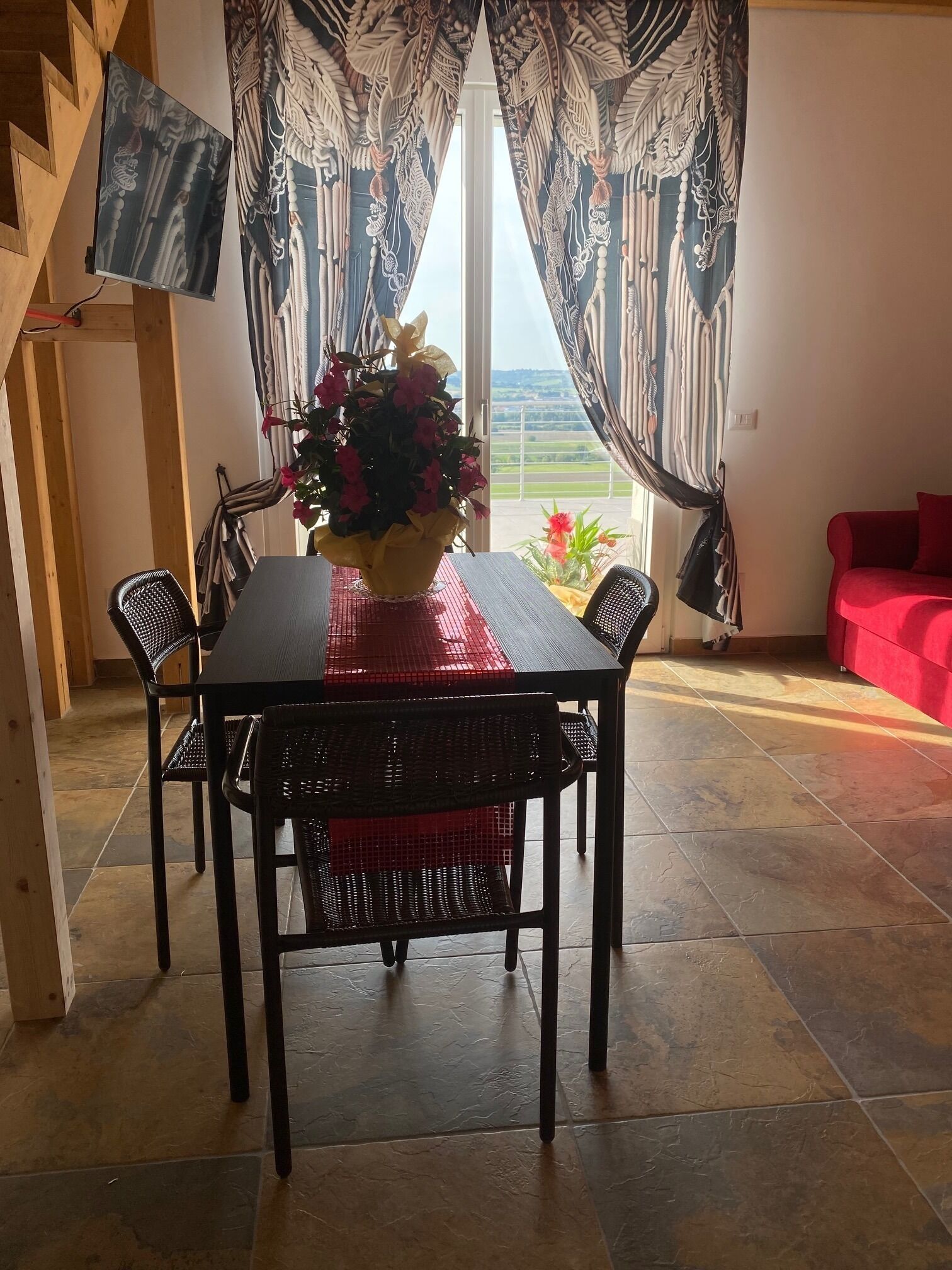 Un comedor con una mesa, sillas, un jarrón de flores y vistas a través de una ventana con cortinas.