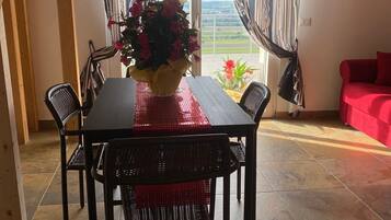 Un comedor con una mesa, sillas, un jarrón de flores y vistas a través de una ventana con cortinas.