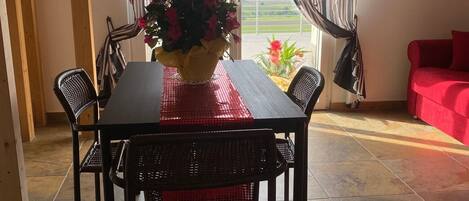 A dining area with a table, chairs, a vase of flowers, and a view through a window with curtains.