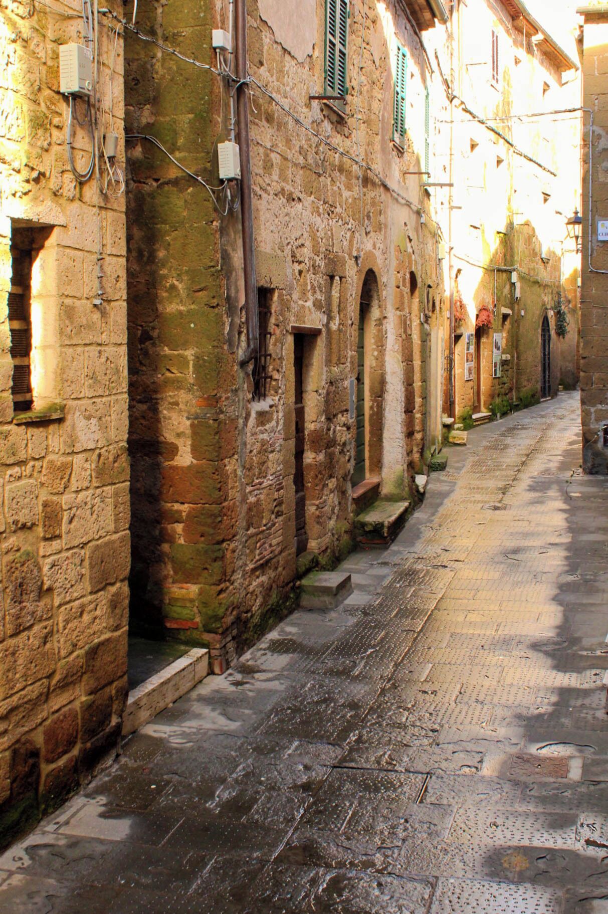 A narrow, cobblestone street lined with historic stone buildings featuring arched doorways and traditional shutters.
