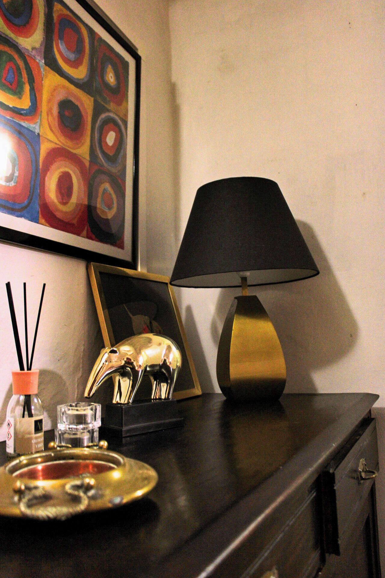 A hotel room with a dark wooden dresser, a black lamp, a framed artwork, and a tray with a bottle and glass.