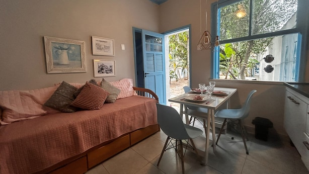 A cozy room with a bed, a dining table, and chairs. There are framed pictures on the wall and a window with a view of greenery outside. - Flor e Fruto Retreat - Little Flower House (Rio de Janeiro)