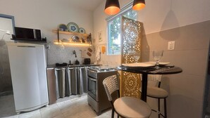 A compact kitchen with a refrigerator, stove, and sink, a small dining area with two stools, and a decorative window panel. - Flor e Fruto Retreat - Little Fruit House (Rio de Janeiro)