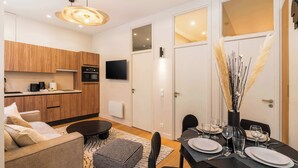 Eine moderne Küche mit Holzausbau, einer schwarzen Arbeitsplatte und einem eingebauten Backofen. Es gibt einen Essbereich mit einem schwarzen Tisch, gedeckt für vier Personen, inklusive Teller, Gläser und Besteck. Ein Fernseher ist an der Wand über der Küche angebracht.