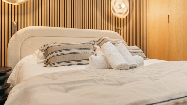 Ein ordentlich bezogenes Bett mit gestreiften und schlichten Kissen, ein runder Nachttisch und eine Wand mit Holzlatten.