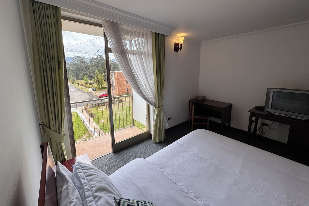 Habitación de hotel con cama, televisión, escritorio y balcón con cortinas.