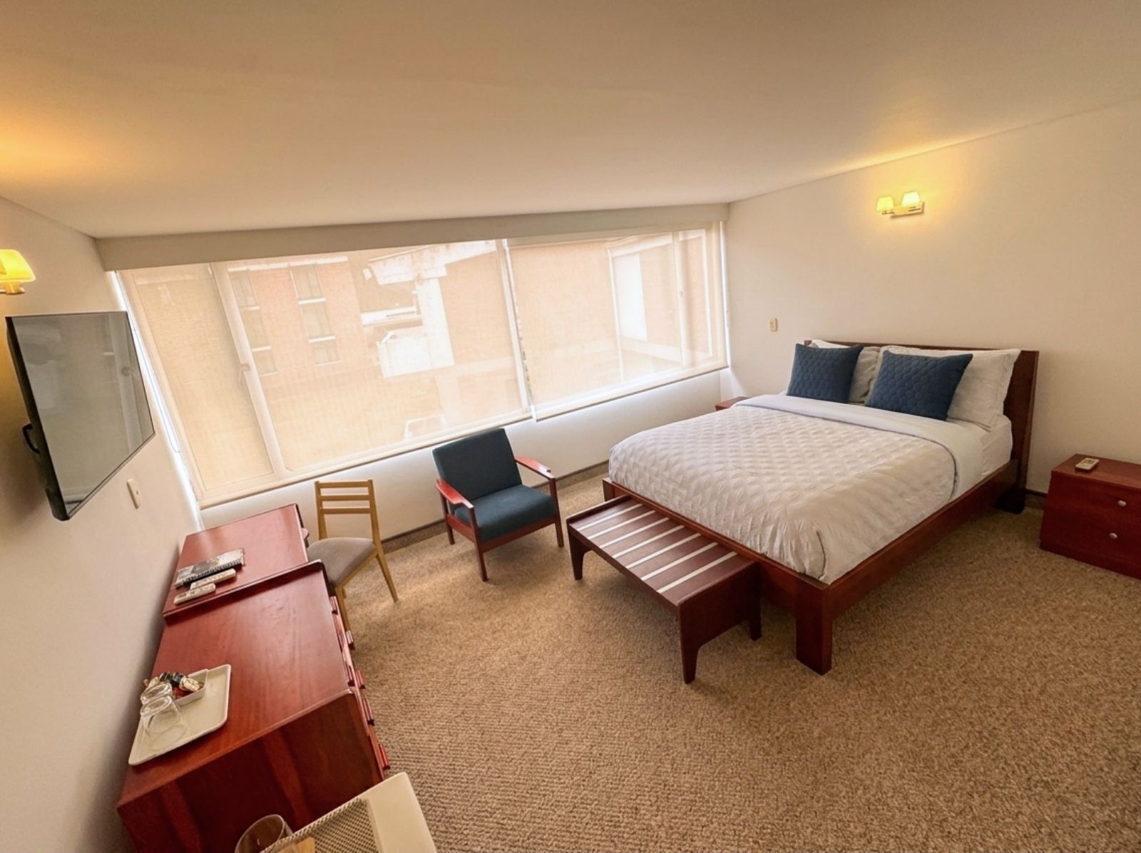 A hotel room with a bed, a desk, a chair, a bench, a TV, and a window with a view.