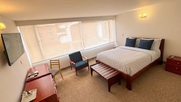 A hotel room with a bed, a desk, a chair, a bench, a TV, and a window with a view.