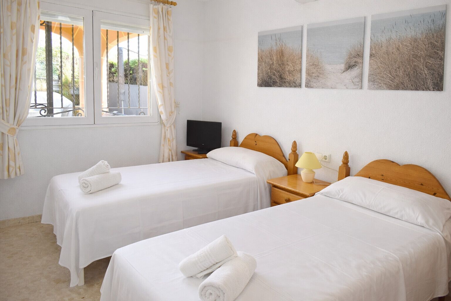 A hotel room with two single beds, a television on a wooden nightstand, and a window with curtains.