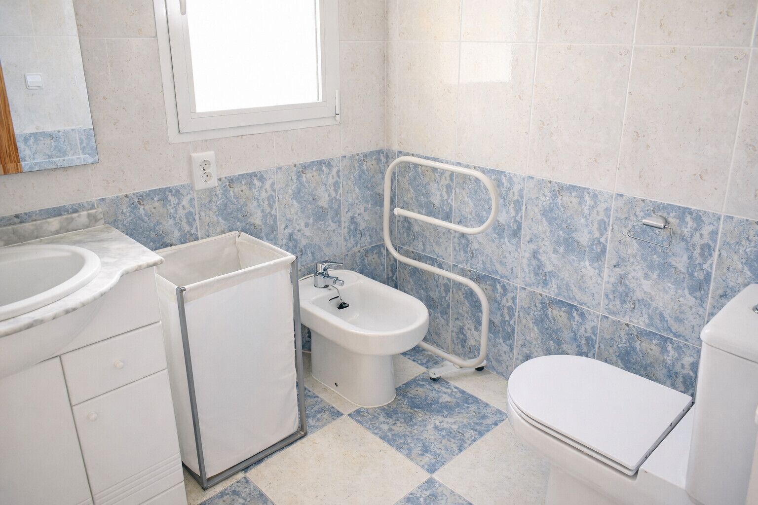 Un baño con lavamanos, bidé y retrete, con azulejos azules y blancos y un ventanal.