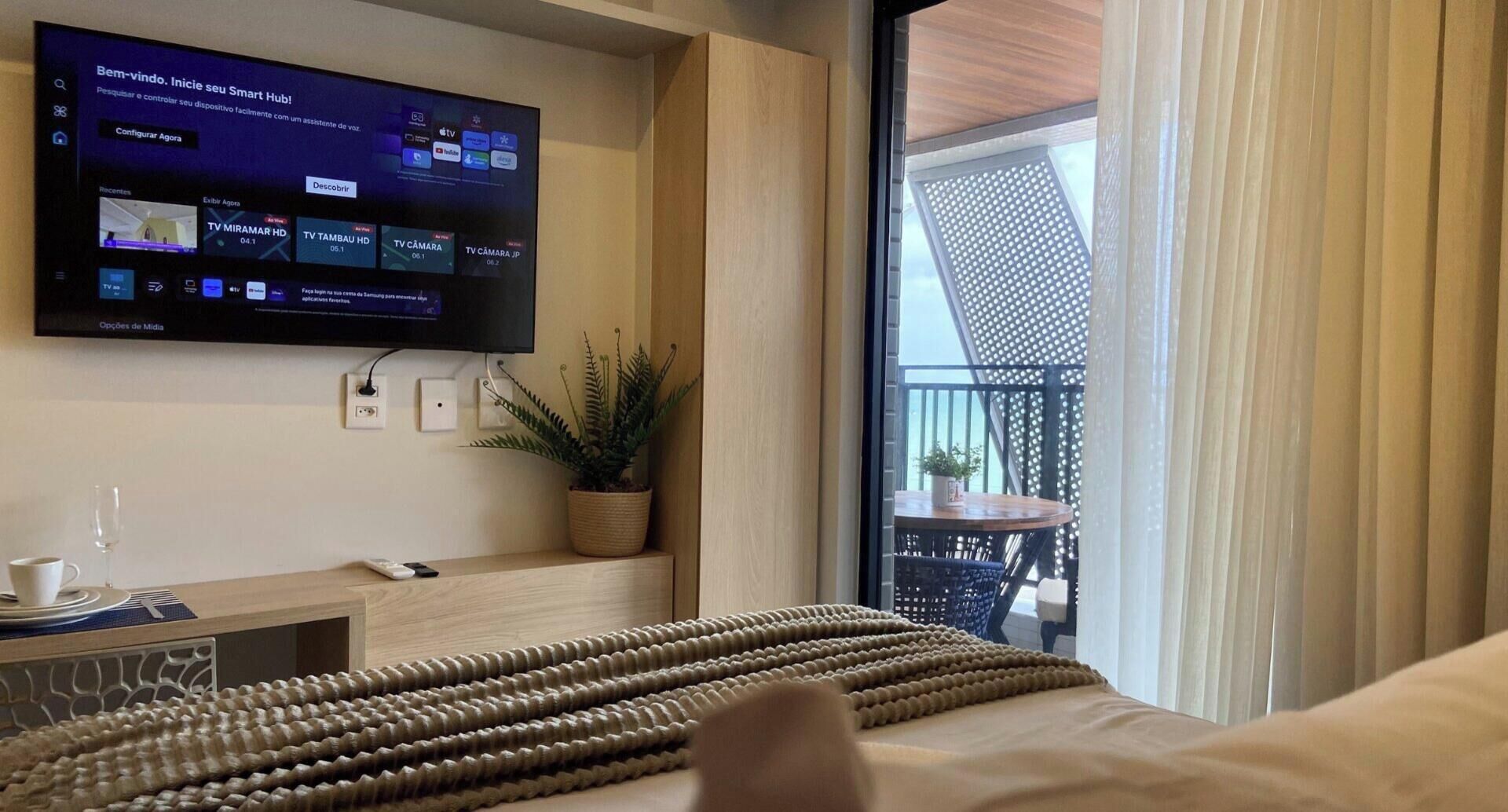 Habitación de hotel con televisor de pantalla plana en la pared, una planta en maceta, una mesita de madera y vistas al balcón.